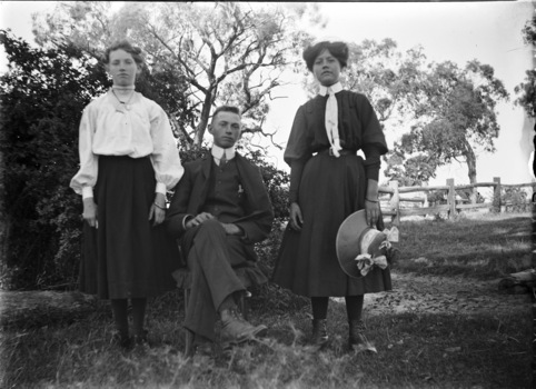 Stan Young seated between two of his sisters who are standing beside him. Trees and a wooden fence are in the background.