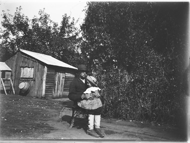 A man seated with a small child on his lap. There is a small wooden building in the background.