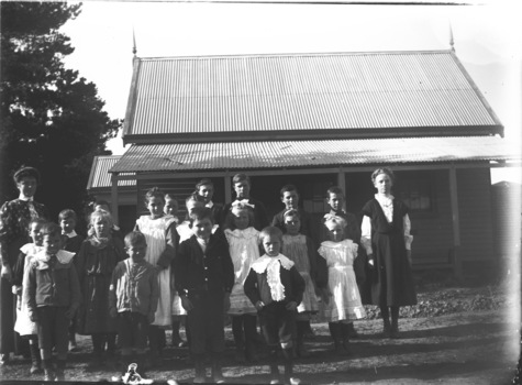 A group of students and their teacher standing in front of the wooden school building,