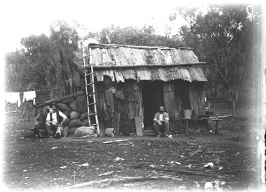 Two men sitting outside a wood and bark slab hut.