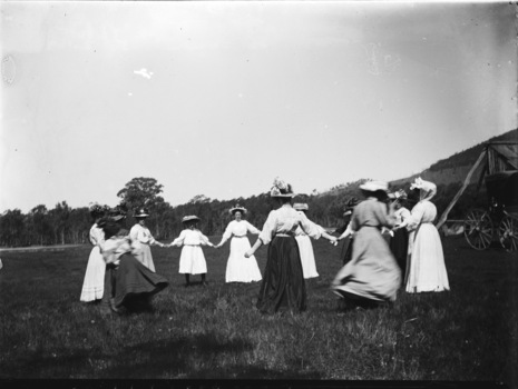 A group of women holding hands in a circle. All are wearing long dresses and hats.