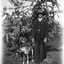 A man and his dog. The man is wearing a three piece suit and a hat. The dog is looking away from the camera. There is a tree in the background.
