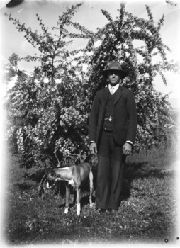 A man and his dog. The man is wearing a three piece suit and a hat. The dog is looking away from the camera. There is a tree in the background.