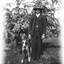 A man and his dog facing toward the camera. The man is wearing a three piece suit and a hat. There is a tree in the background.