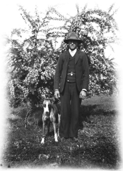 A man and his dog facing toward the camera. The man is wearing a three piece suit and a hat. There is a tree in the background.