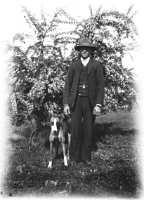 A man and his dog facing toward the camera. The man is wearing a three piece suit and a hat. There is a tree in the background.
