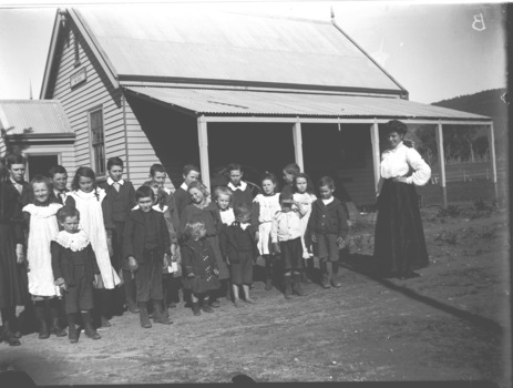 A group of students standing outside their school building with the teacher on the right of the image.