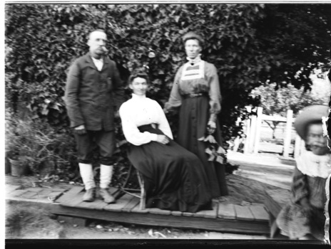 A man, two women and a child seated in front of a tree. A child is on the right hand edge of the image.