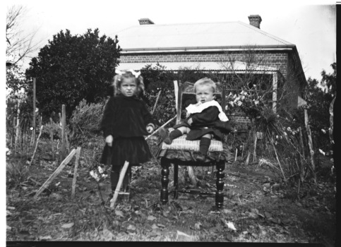 A young girl standing beside a younger child seated on a chair. Their brick house is in the background.