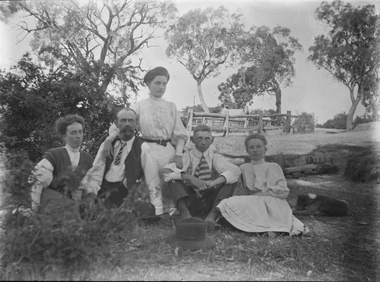 A family group of husband, wife and three children seated outside. A house and fenced yard is in the background.