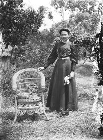 A woman standing beside a cane chair. One hand on the back of the chair, the other holding a flower.