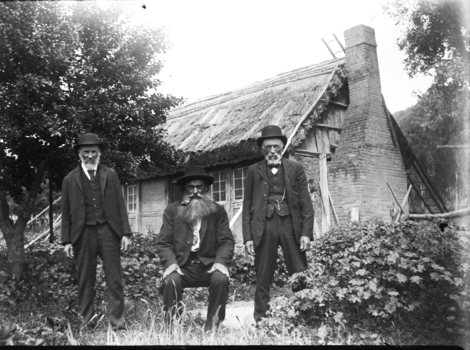 Two men standing and another seated between them.  A house is in the background.