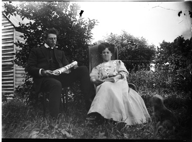 A man and woman seated in a garden. He is smoking a pipe and reading the newspaper.