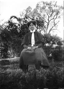 An unidentified woman seated on a chair outside. There is a house partially hidden in the trees on the right of the scene.