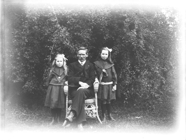 A man seated on a chair between two girls. There are trees in the background.