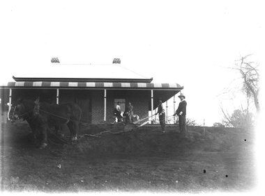 Two men ploughing with a pair of horses. A couple with dogs are on the verandah in the background.