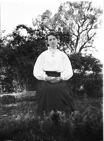 A young woman seated outside. She is wearing a white, high-necked blouse and dark skirt.