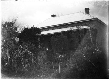 A man a dog standing on a verandah. The house is made of brick with a roof of corrugated iron.