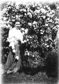A woman standing in front of a flowering tree. She is wearing a long-sleeved white blouse and long, dark coloured skirt.