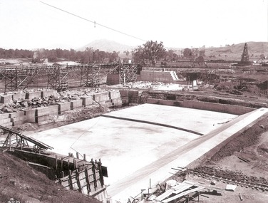 A stilling pool in foreground being constructed on the downstream side of a dam.