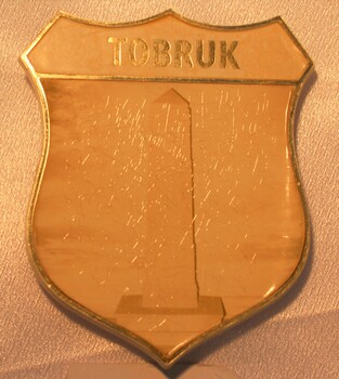 Small plastic shield with Tobruk written in gold and the Tobruk Memorial in the centre.