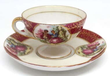 A small cup and saucer with image of a couple in a garden and red and gold design