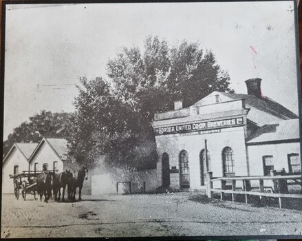 Black and white photo of brewery buildings. A horse drawn cart is in the left foreground.