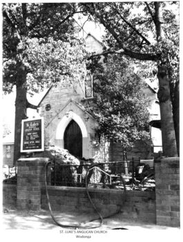 Later photo of St Luke's church with notice board showing service times. A hose is draped over the fence.