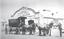 A coach pulled by 4 horses in front of the Federal Coach Factory. 4 men including 2 Indian hawkers are in the foreground.