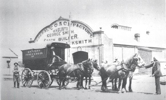 A coach pulled by 4 horses in front of the Federal Coach Factory. 4 men including 2 Indian hawkers are in the foreground.