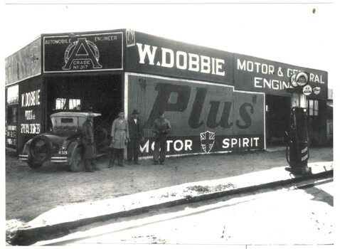 4 men and a car standing outside engineering workshop. A petrol bowser is on the right foreground.