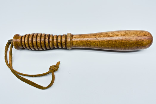 A golden coloured small wooden truncheon that features a grooved handle and a leather wrist strap.