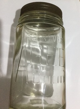 A cylindrical, tall clear glass Agee preserving jar with a tin lid.