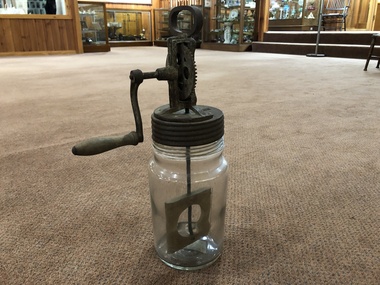 A tall glass butter churn with a metal screw top lid, a metal handle to hold it steady and a turning mechanism on the top.   