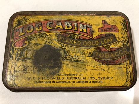 A small rectangular, metal 'Log Cabin' tobacco tin with red edging around the yellow, hinged lid.