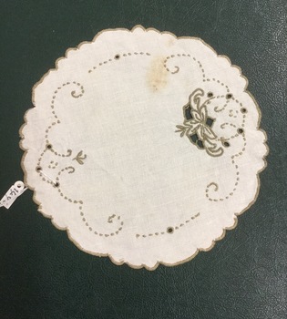 A round cream linen doily with khaki coloured floral embroidery, open cut pattern and a scalloped edge.