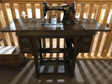 A 'Free' brand 1920's treadle sewing machine with ornate gold decals on it. It is in a wooden sewing cabinet with four drawers at the front.