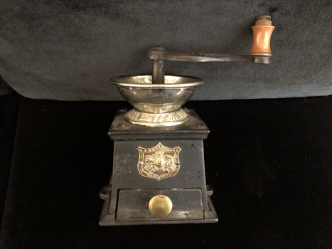 A vintage metal and brass coffee grinder with a curved shaped turning handle which has a turned wooden knob at the end. 