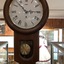A brown wooden wall clock with a white round clock face, with roman numerals, and a brass pendulum.