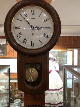 A brown wooden wall clock with a white round clock face, with roman numerals, and a brass pendulum.