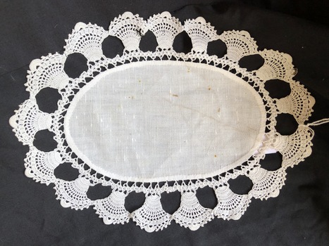 An oval shaped white cotton doily with a wide fluted shaped patterned crocheted edge.
