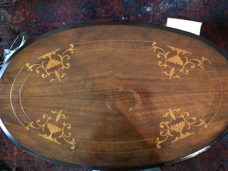 An antique brown oval table with inset gold coloured wooden ornate urns and a double lined pattern around the edge on the top.