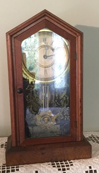 A wooden steeple cased mantel clock with a hinged highly decorative glass front, black roman numerals and metal hands on the enclosed white face.