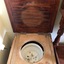 A brown stained wooden commode with short legs on wheels. It has an enamel white chamber pot under the lid.