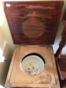 A brown stained wooden commode with short legs on wheels. It has an enamel white chamber pot under the lid.