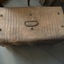 A brown damaged metal trunk with a hinged lid and metal handles on the sides and top. It has brown or red paint marks. There are six studs on the lid.