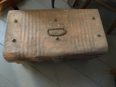 A brown damaged metal trunk with a hinged lid and metal handles on the sides and top. It has brown or red paint marks. There are six studs on the lid.