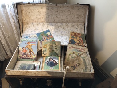 A large leather handled travelling cabin trunk with a hinged lid. It contains six books and a skipping rope.