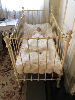 A white painted iron cot with drop down sides and castors to move it.  Large knobs are on the top and bottom of the rungs. 