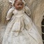 A porcelain or ceramic baby doll dressed in a long white cotton and lace dress laying on a white crocheted rug.